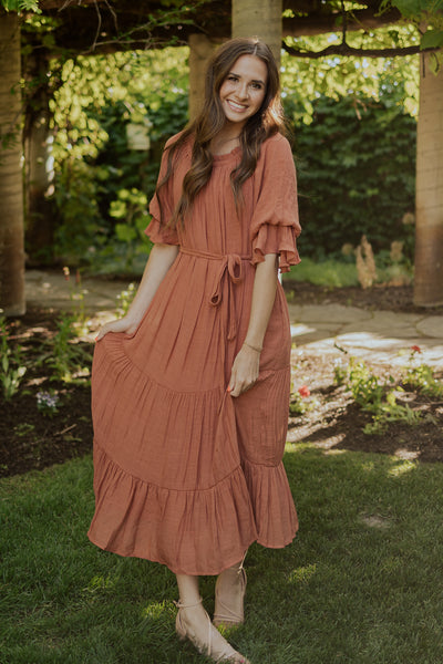 A front shot of a rusty, orange, modest dress featuring a belt, scoop neckline, and fun, puff sleeves.