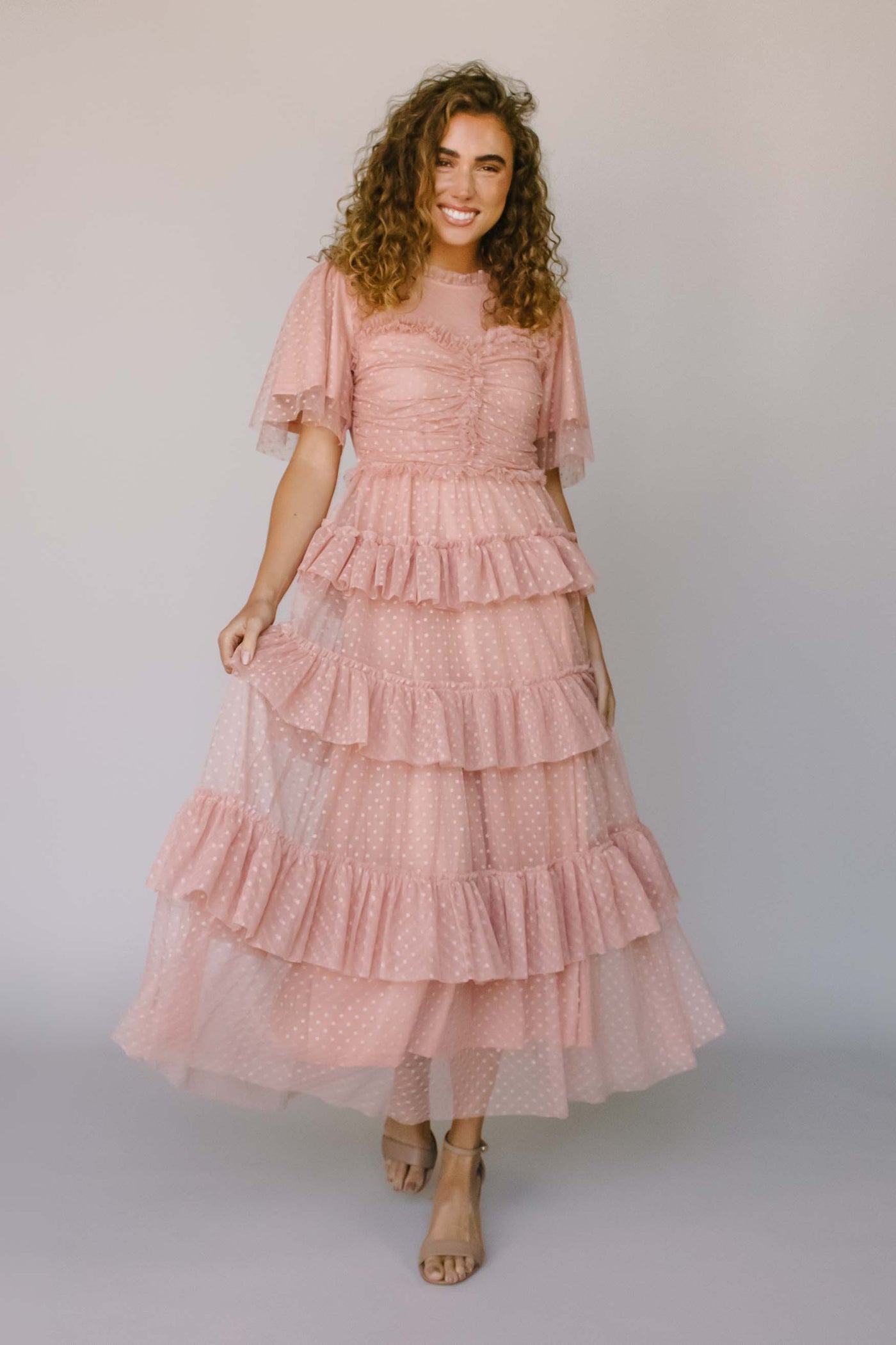 A modest dress in the color blush pink with a fun dotted fabric, layered tiers, and flutter sleeves. It also features a fun smocked pattern on the bodice and a high neckline.