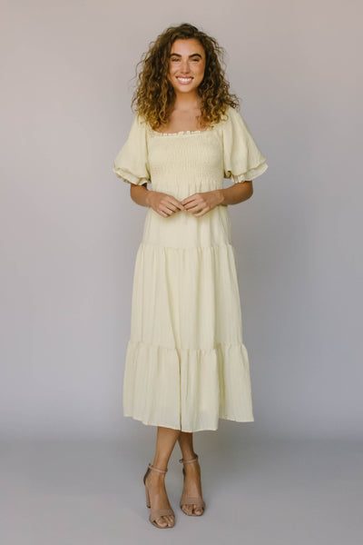A yellow modest dress with a square neckline, ruffled puff sleeves, smocked bodice, and tiers.
