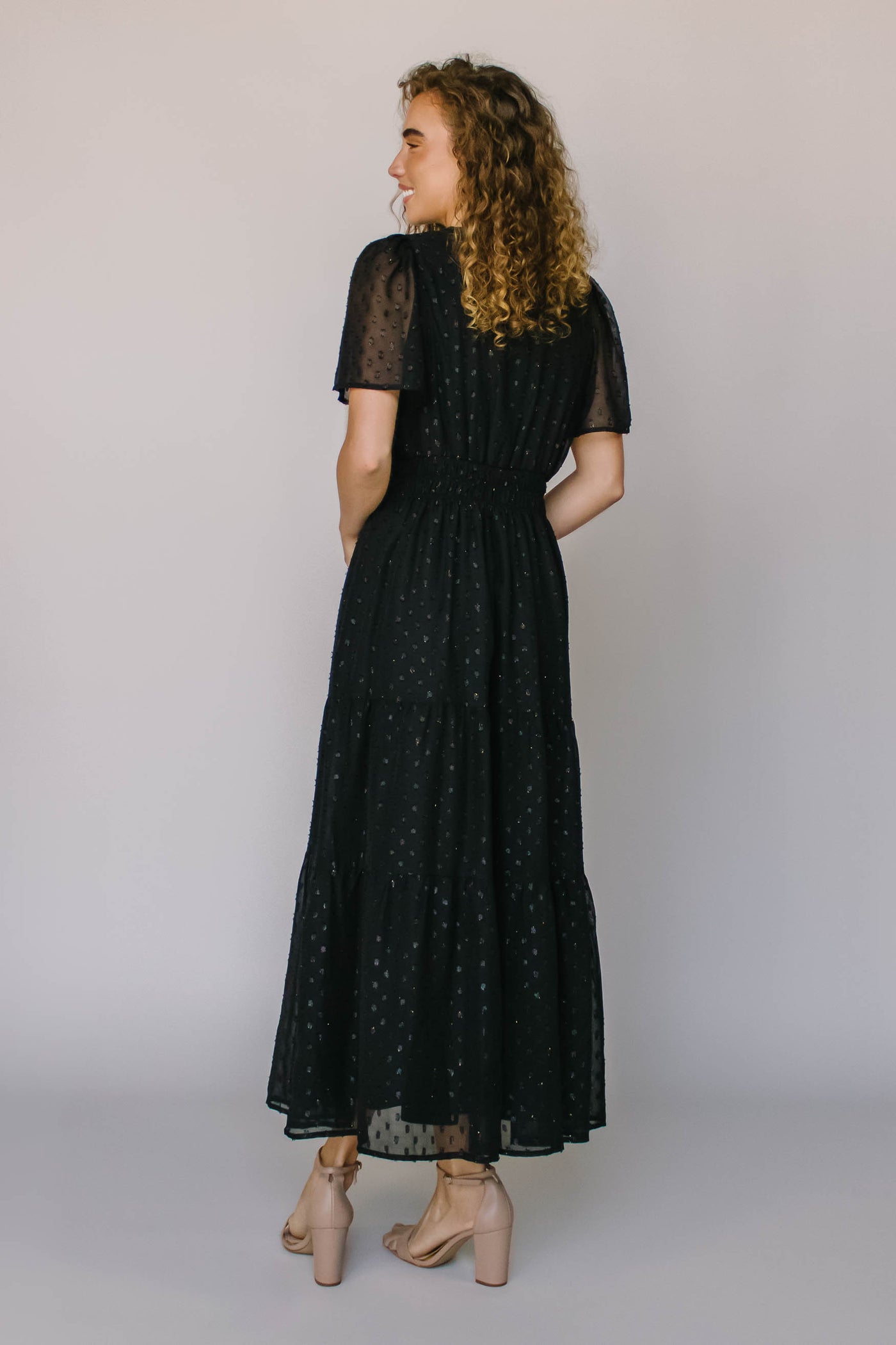 The back of a modest dress in black with a shimmery black spotted fabric, a defined waist, and flutter sleeves.