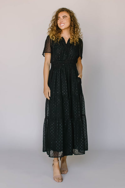 A modest dress in black with a shimmery black spotted fabric, a defined waist, and flutter sleeves.
