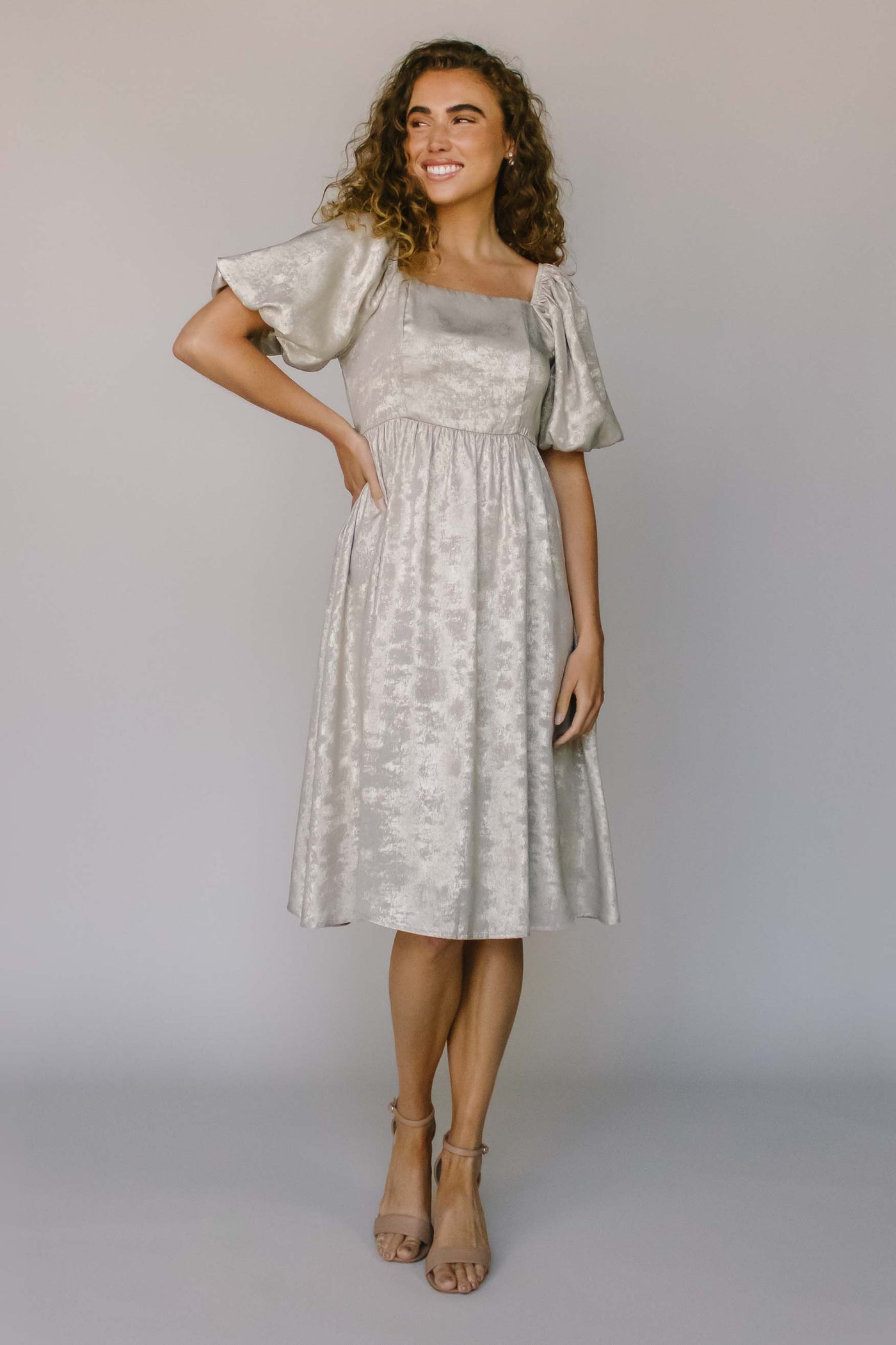 A modest dress in a shimmery champagne color with puff sleeves, a defined waistline, and a stunning square neckline.