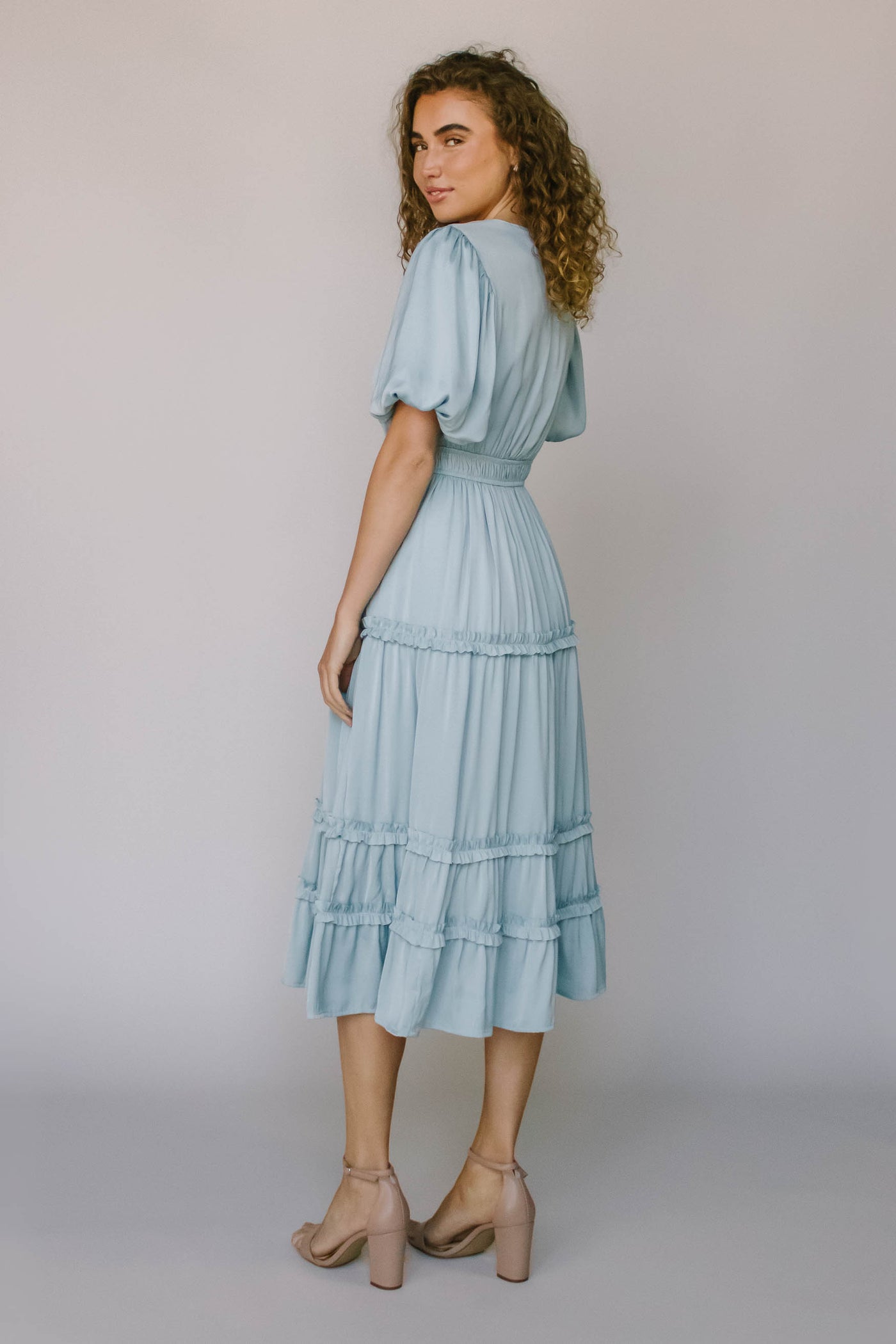 The back of a modest dress in French blue with puff sleeves, ruffled tiers, a defined waistline, and puff sleeves.