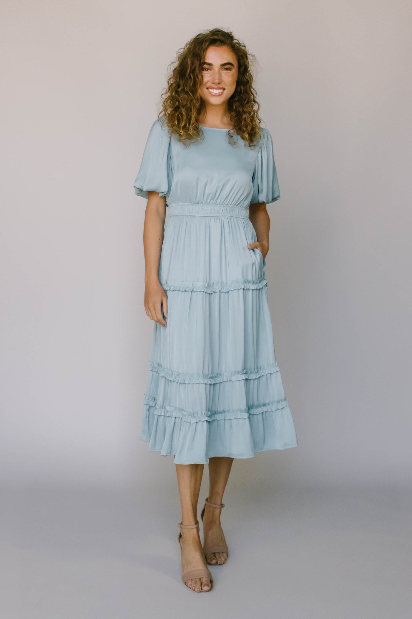 A modest bridesmaid dress in French blue with ruffled tiers, defined waist, puff sleeves, and a high neckline.