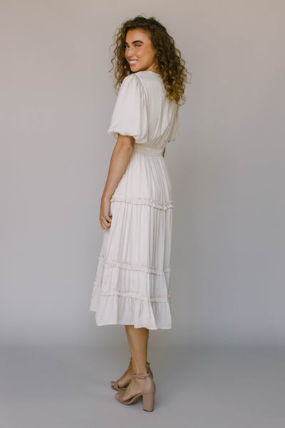 The back of a modest dress in ivory with puff sleeves, ruffled tiers, a defined waistline, and puff sleeves.