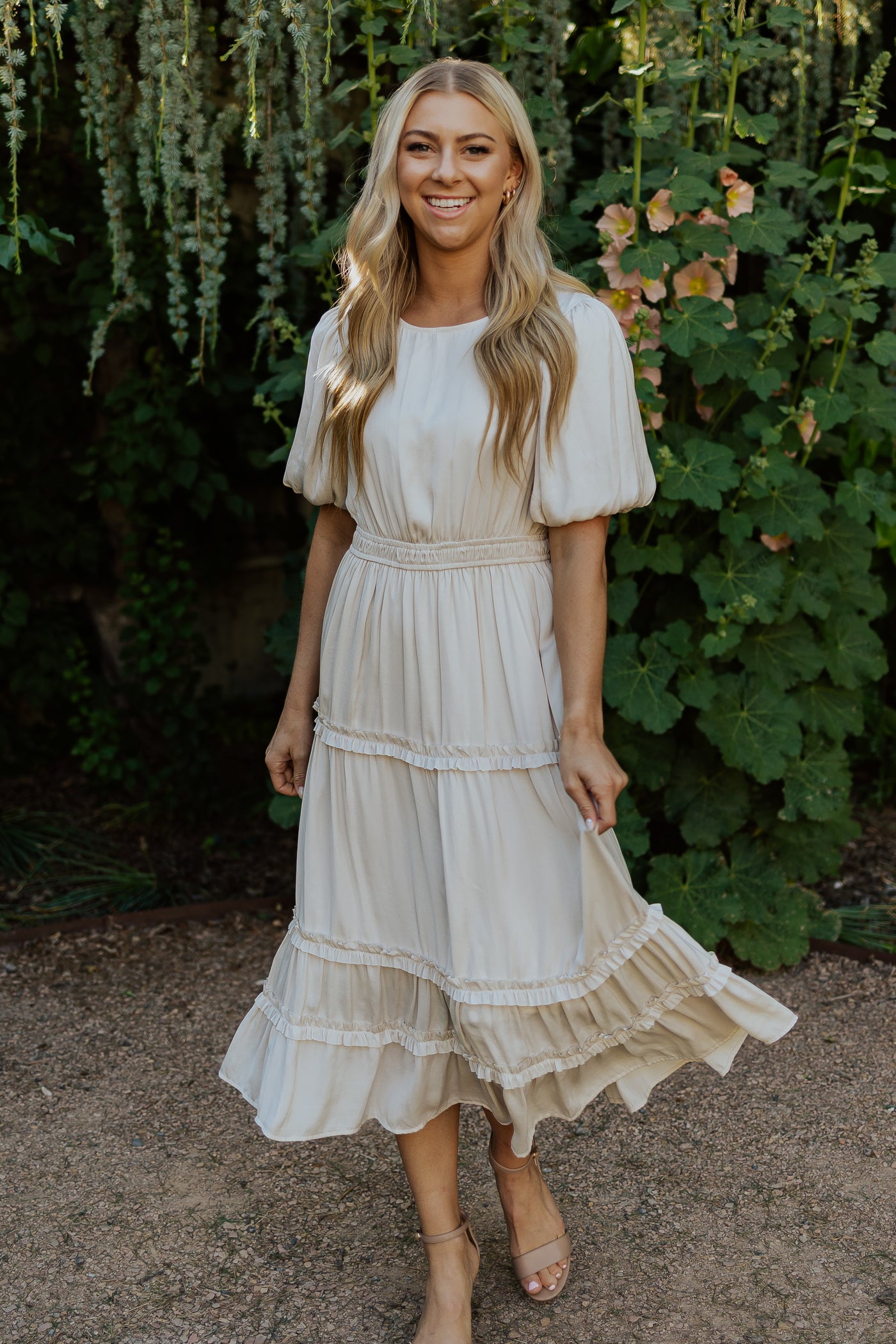 An alternate shot of the front of a cream, modest dress. The dress features a midi length skirt, defined waistline, and billowy sleeves.