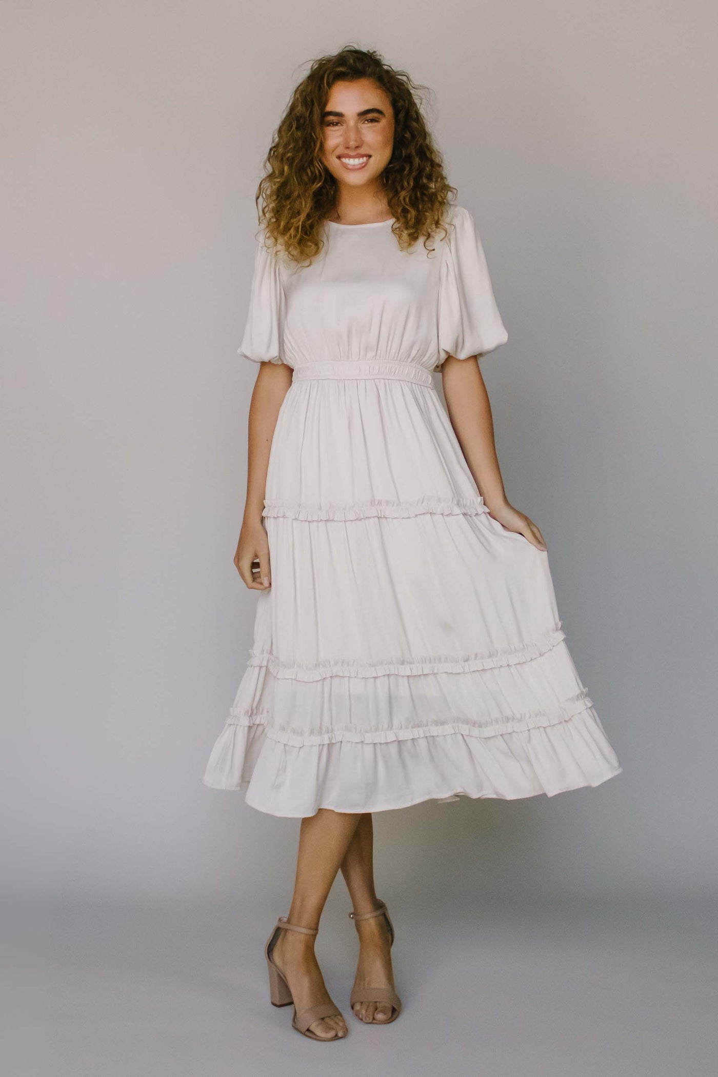 A modest dress in ivory with ruffled tiers, defined waist, puff sleeves, and a high neckline.