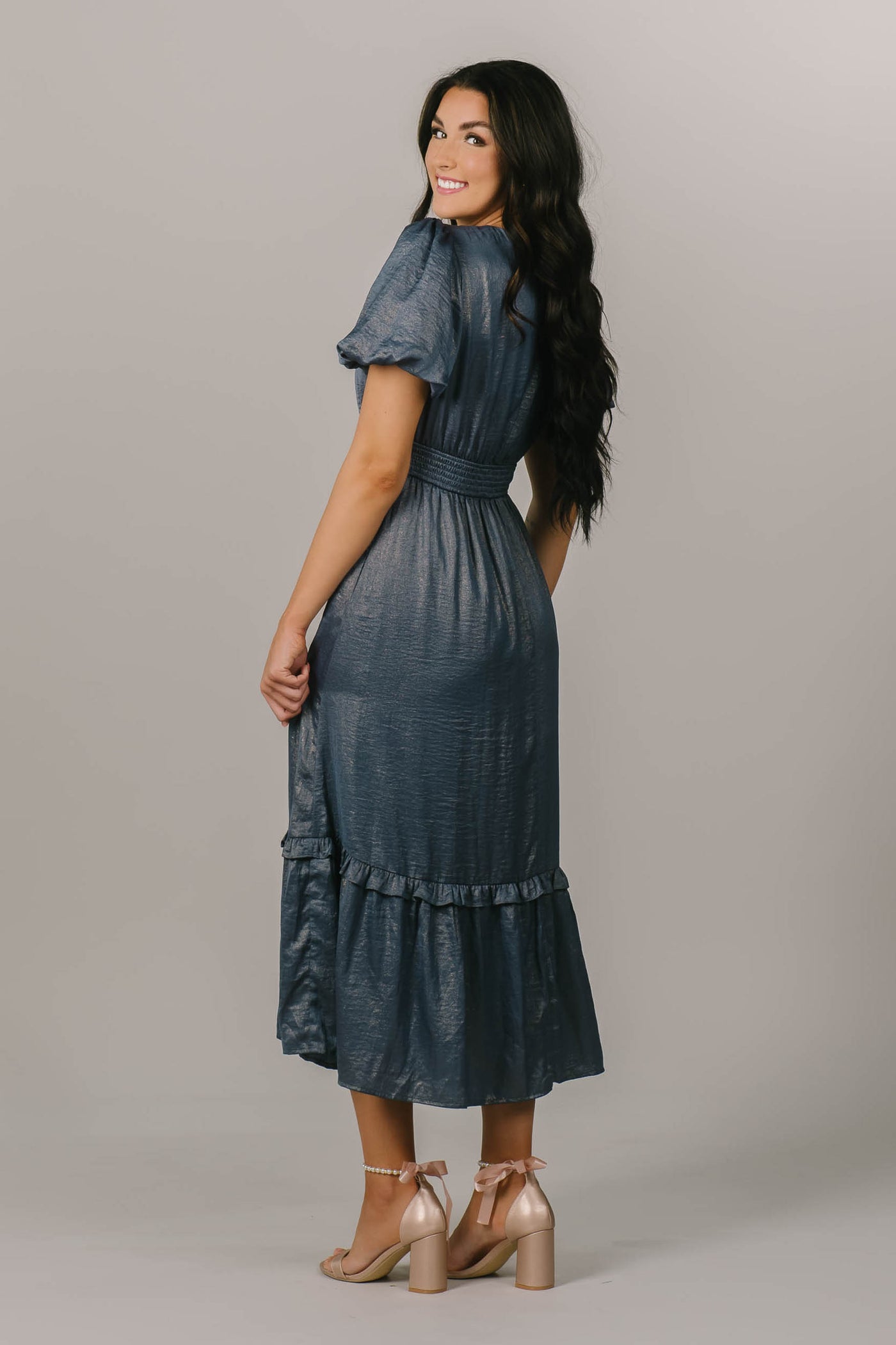 This a brunette model wearing a modest dress with shimmery blue fabric and a shot puff sleeve.