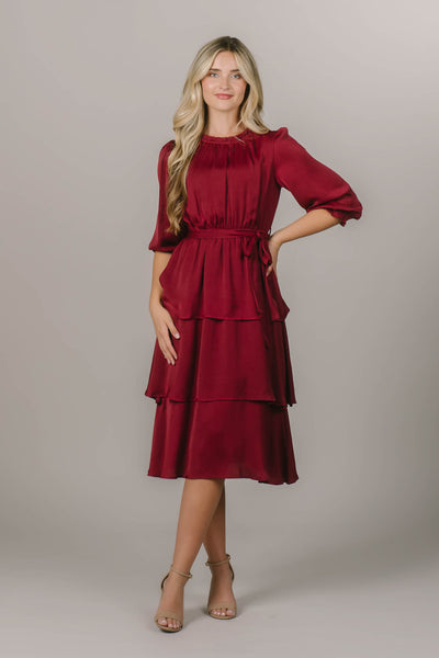This is a modest dress featuring a red color, tiered skirt and waist tie.
