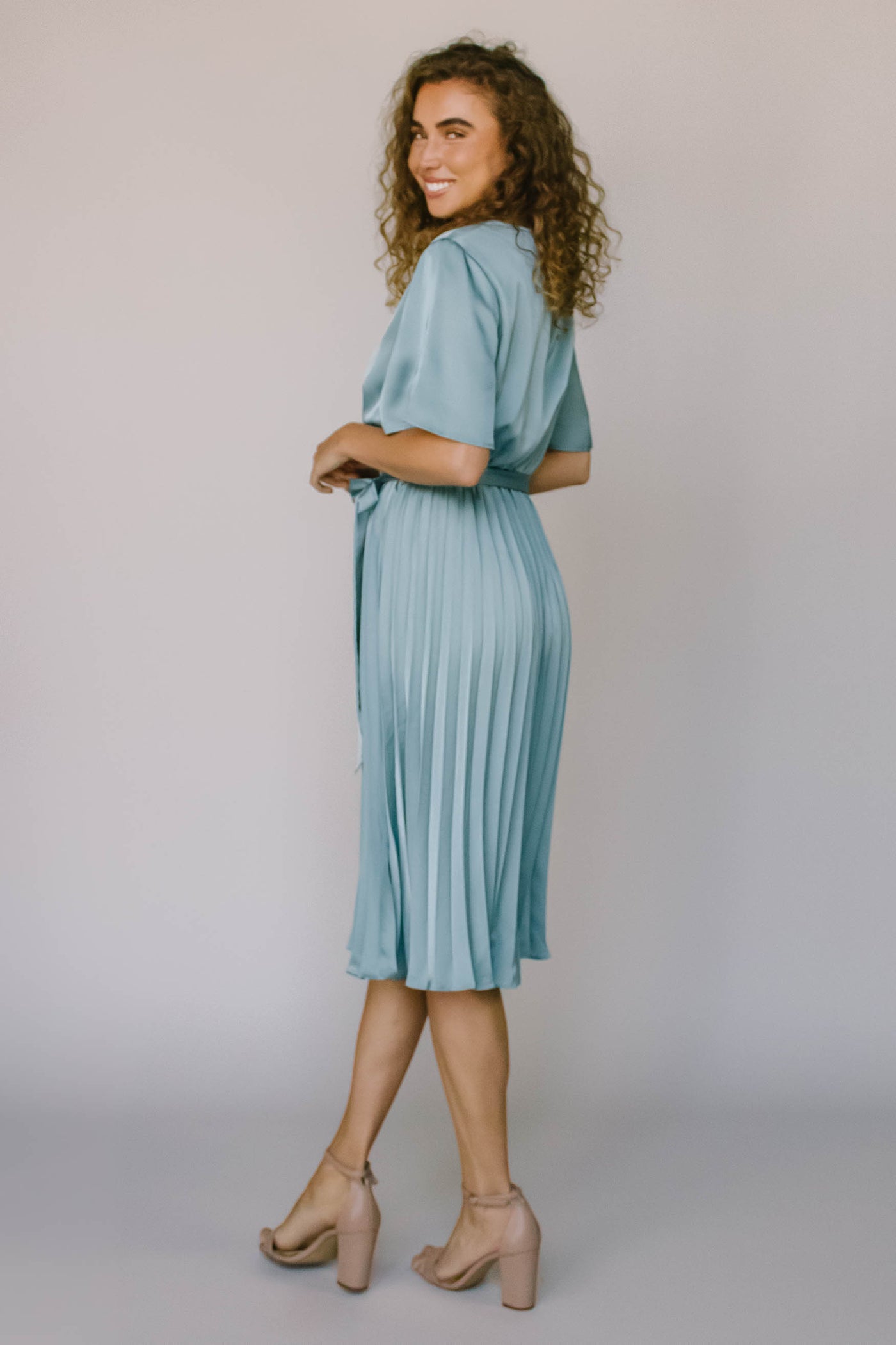 The back of a modest dress in a blue silky fabric, flutter sleeves, a pleated skirts, and a defined waist with a tie.