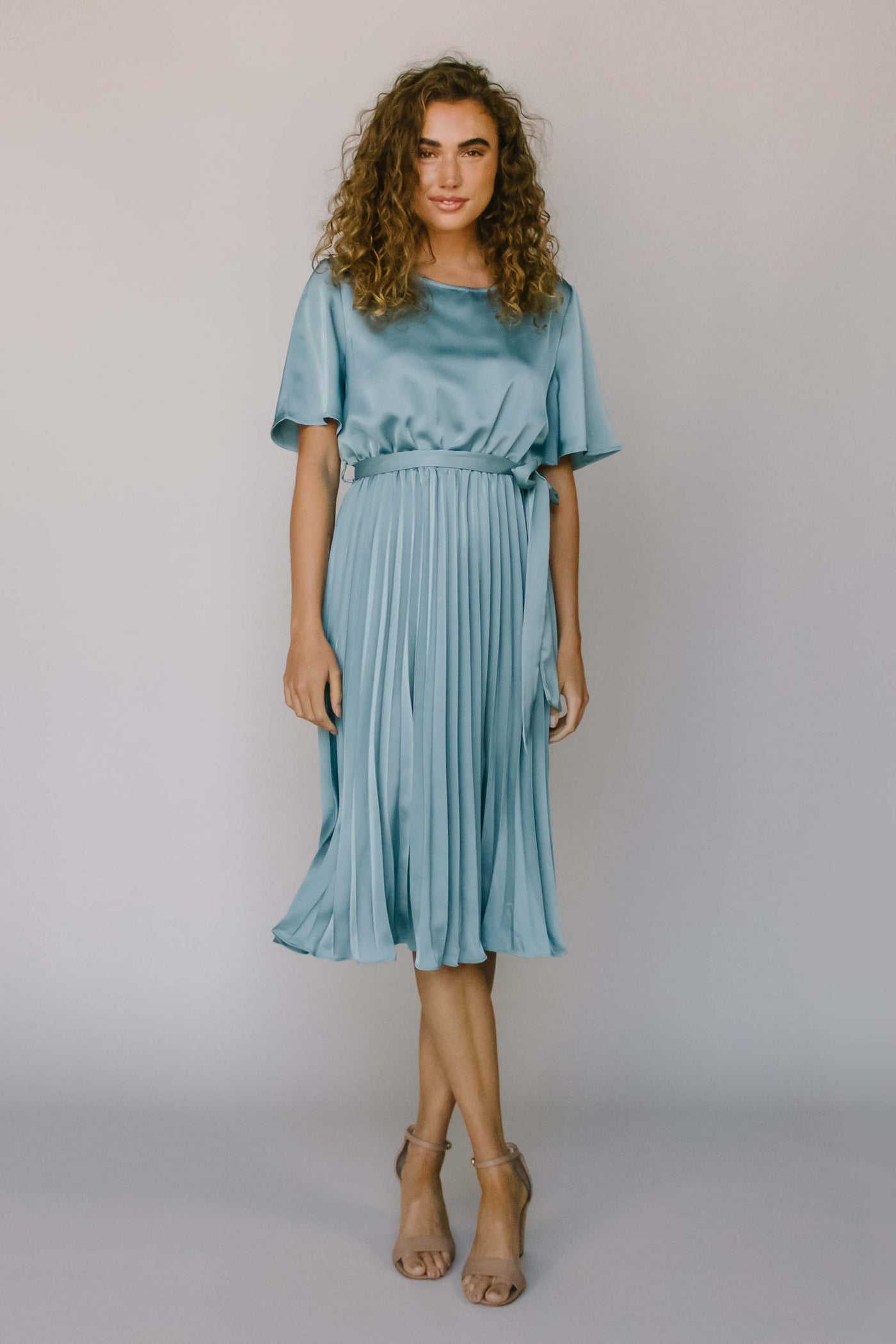 A modest dress in a silky blue fabric with flutter sleeves, a high neckline, a pleated skirt, and a defined waist with a bow.