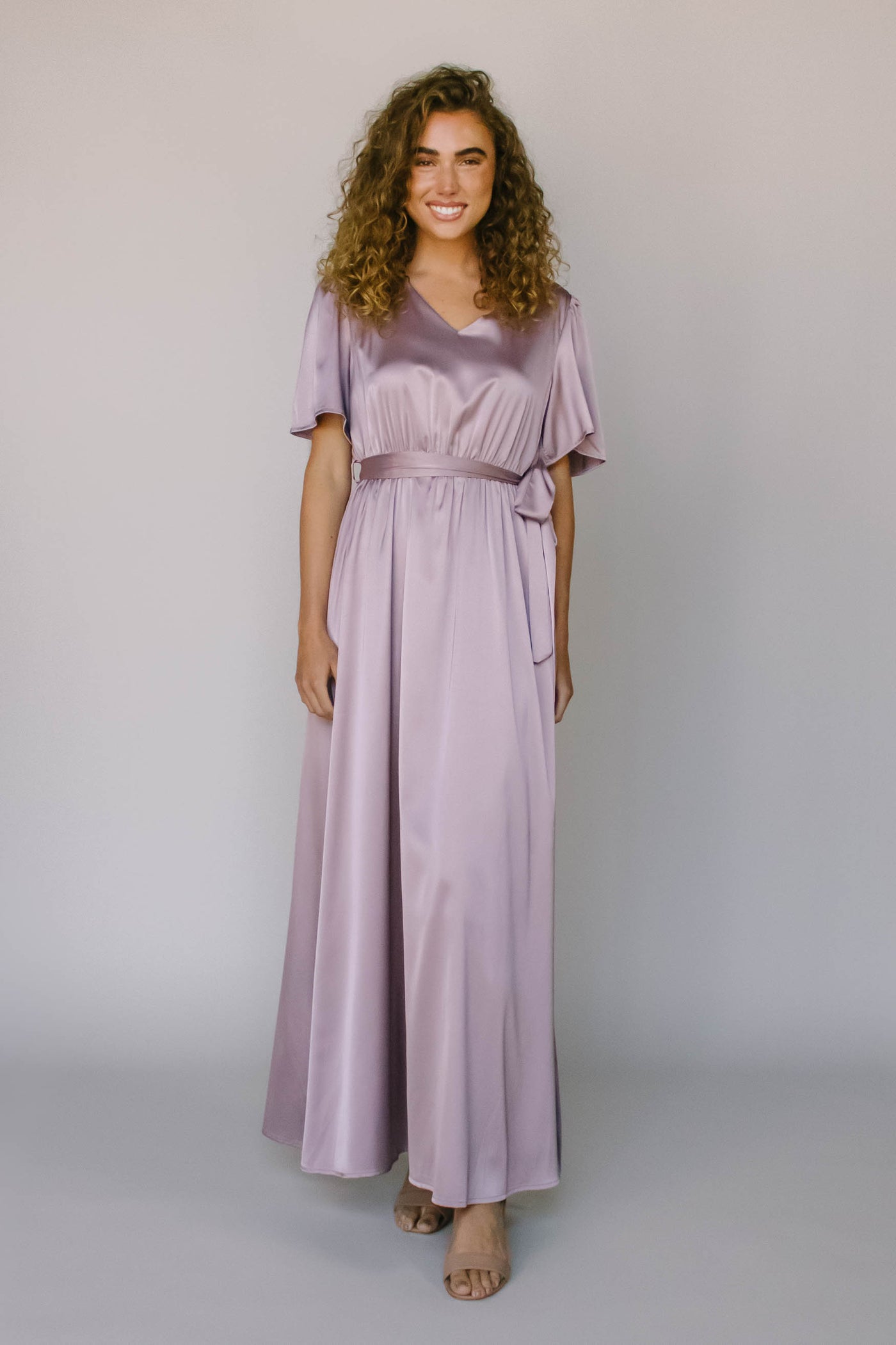 A full length modest bridesmaid dress with a silky purple fabric, flutter sleeves, v-neckline, and a tied bow on the waist.