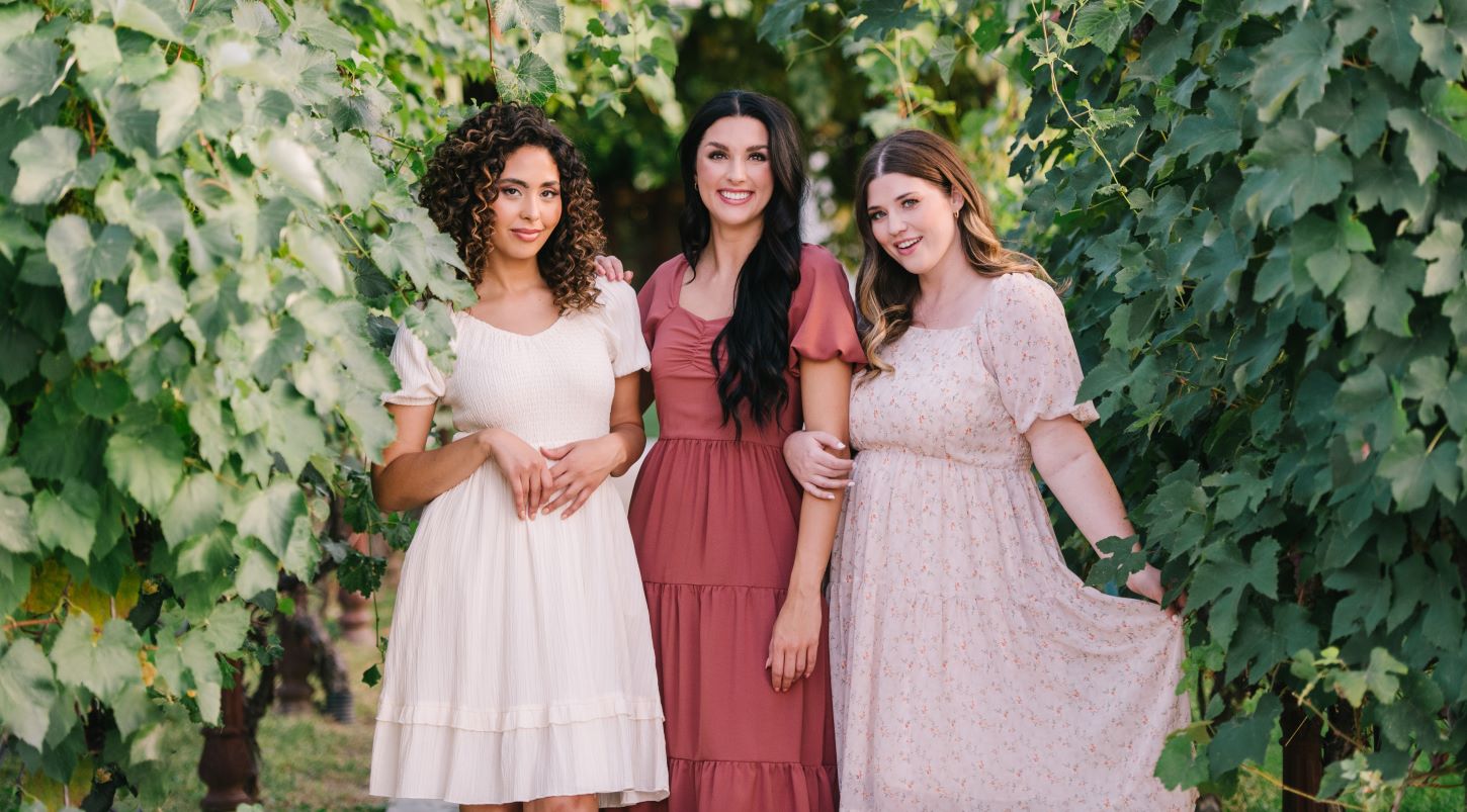 Three ladies standing in a vineyard wearing modest dresses in different colors