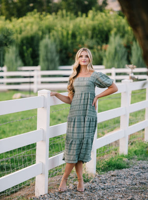 A woman wearing a sage green modest dress with a textured pattern leaning against a white picket fence