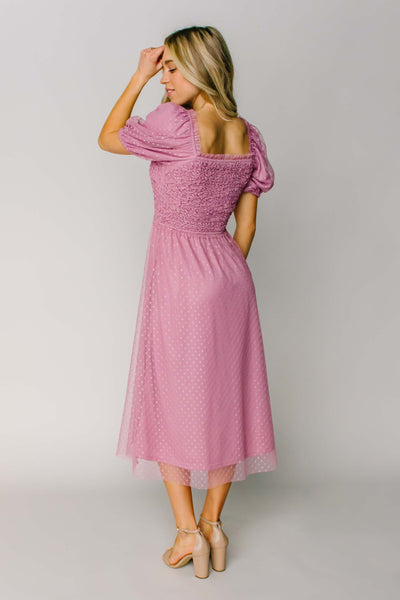 The back of a modest dress in a pink color with a square back neckline, puff sleeves, and a swiss dot fabric.