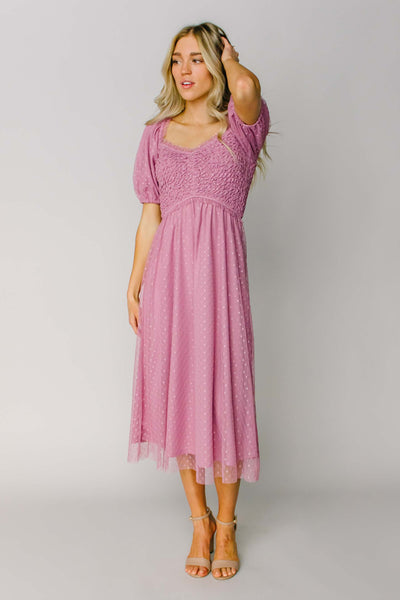A modest dress in a pink color featuring a square v-neckline, puff sleeves, a smocked bodice, and swiss dot fabric.