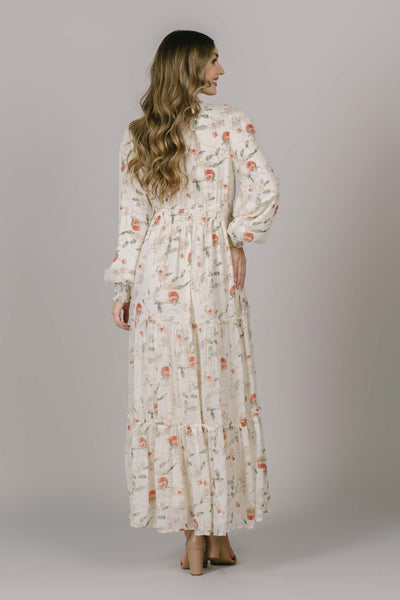 The back of a full length modest dress with a light peach pattern, bishop sleeves, tiers, and a defined waistline.