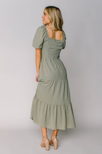 The back of a modest dress in a sage green color with a square back neckline, puff sleeves, and tiers.