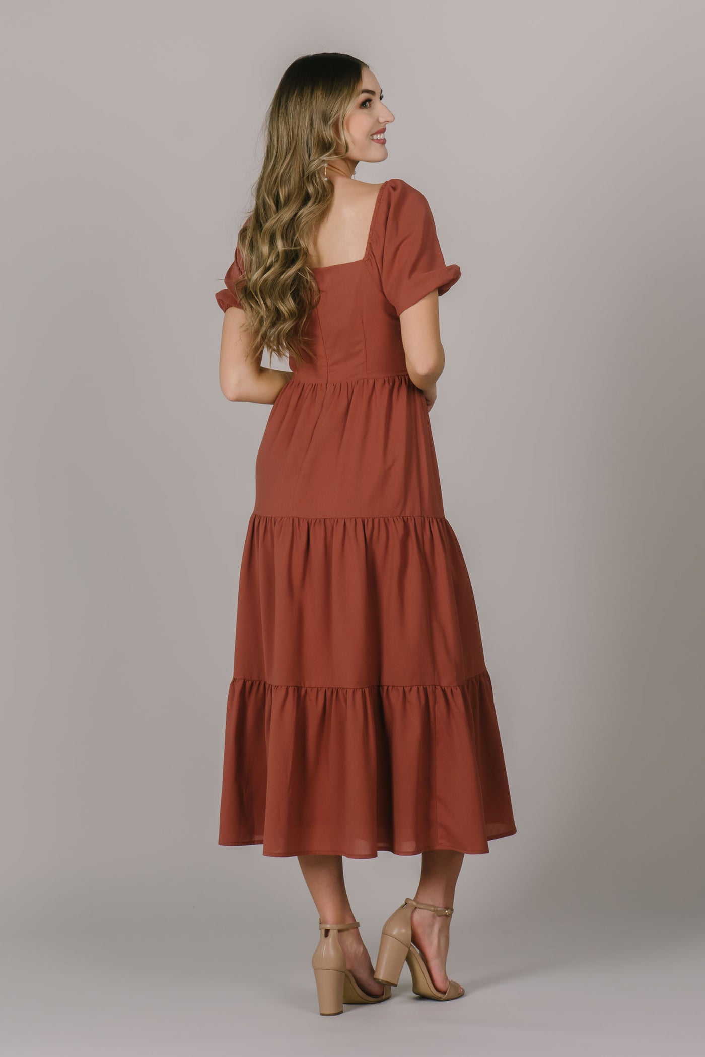 The back of a modest dress in a rust color with a square back neckline, puff sleeves, and tiers.