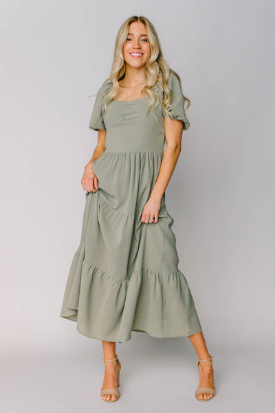A modest dress in a beautiful sage green color with a square neckline, puff sleeves, and a rouched bustline.