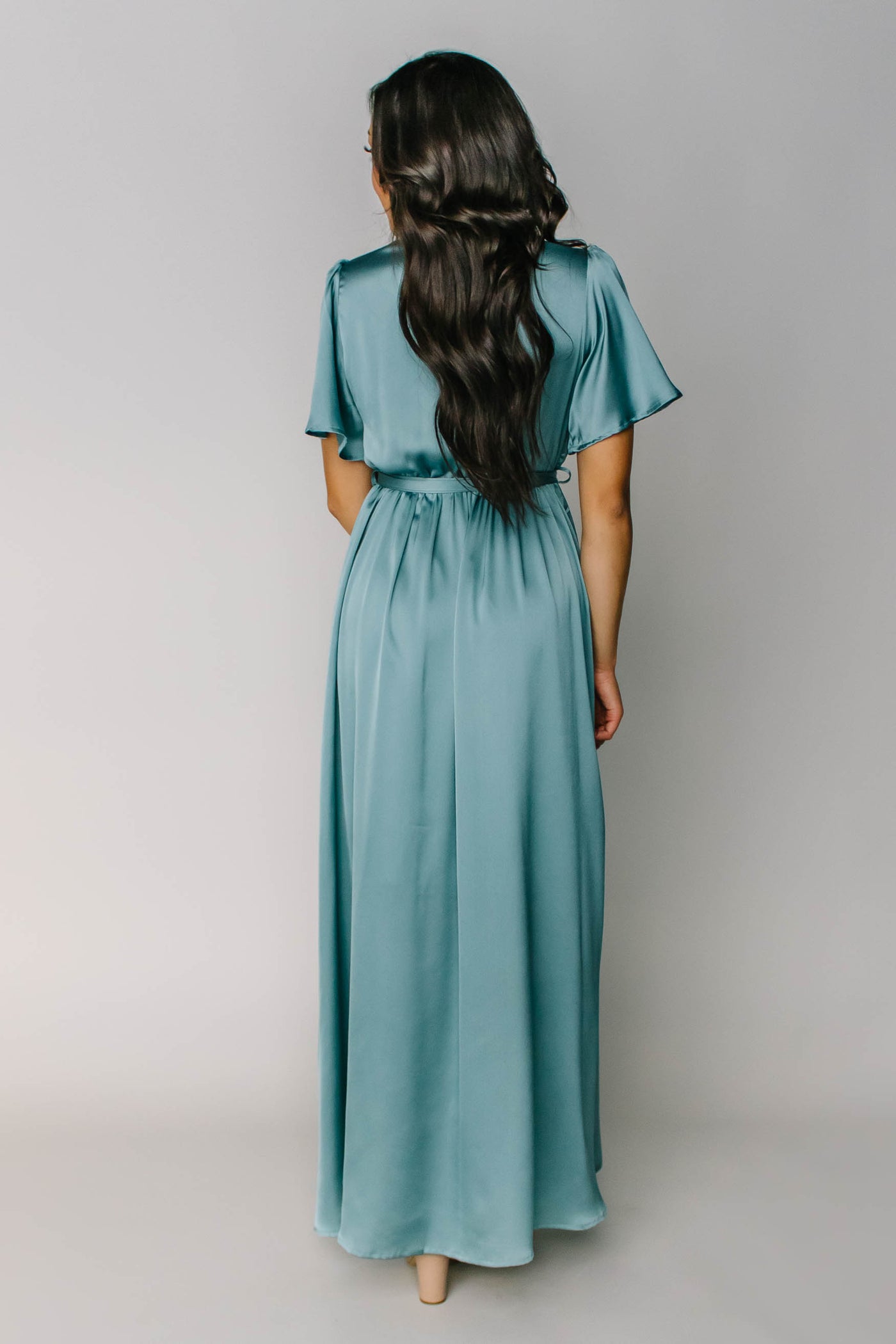 The back of a full length modest bridesmaid dress in a teal blue color with a tied waist and flutter sleeves.