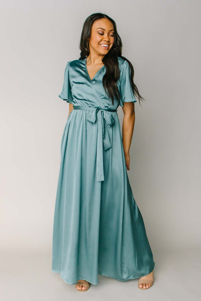 A modest bridemaid dress in a beautiful teal blue color, made with a silky soft material. This dress features a mock wrapped bodice, a tied bow, and flutter sleeves.
