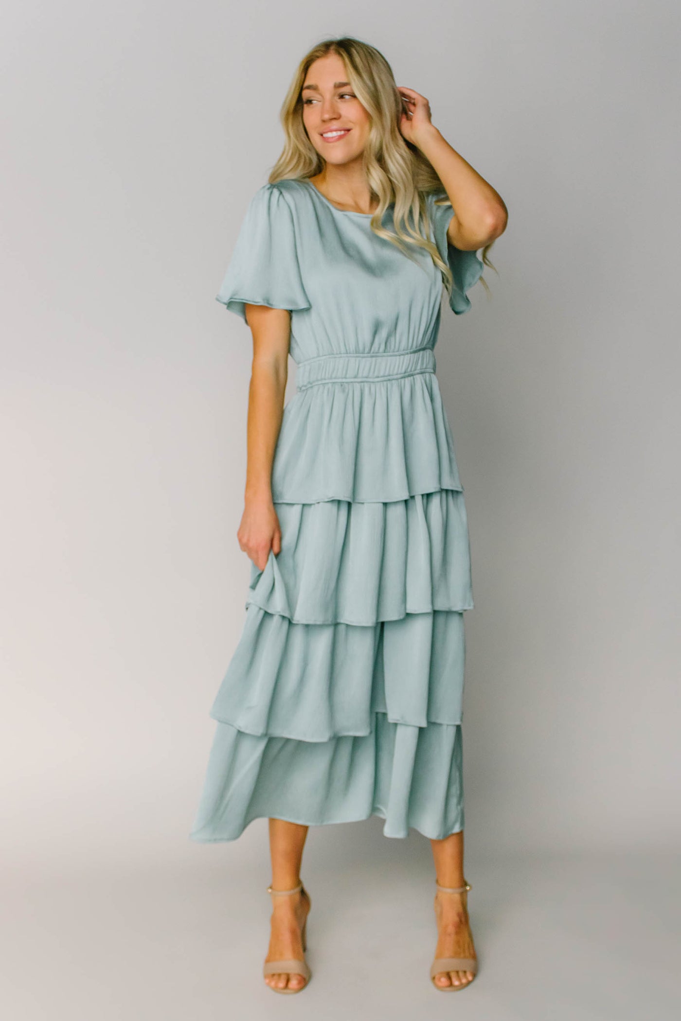 A modest bridesmaid dress made out of a silky light blue fabric with flutter sleeves, layered tiers, and a defined waist.