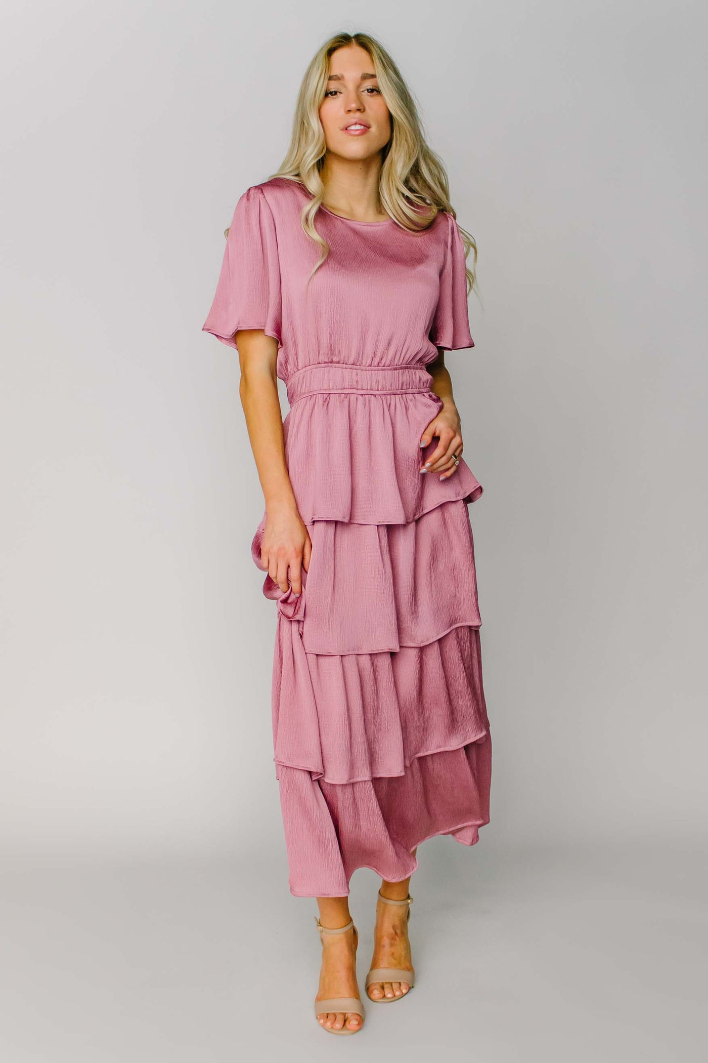 A modest bridesmaid dress made out of a silky pink fabric with flutter sleeves, layered tiers, and a defined waist.