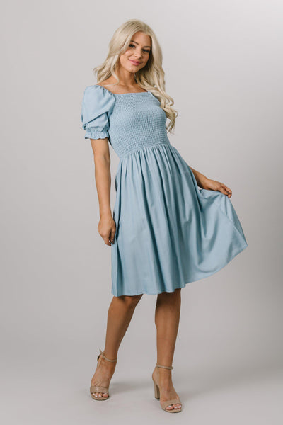 Modest Dresses -  Everyday Modest Dresses - Bridesmaid Modest Dresses. This model is wearing a small knee length dress thah has a smocked top and flowy skirt. It features a square neckline with puff sleeves, the dress is faded denim sheen and from a dress shop called Moments Made.