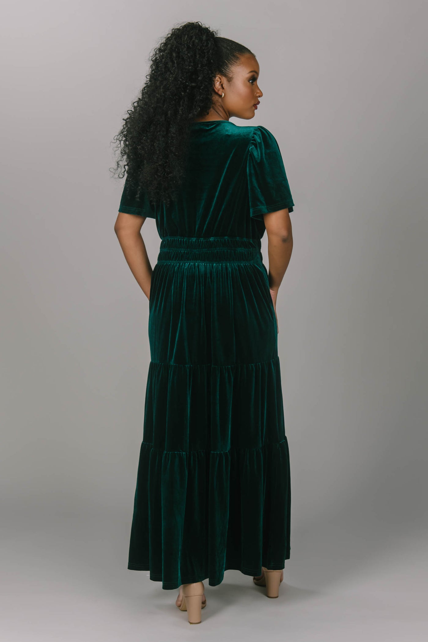 Back view of modest everyday and bridesmaid emerald colored dress. This dress is velvet, has a v-neckline, and a tiered skirt. This modest dress for women is floor length and super comfortable.