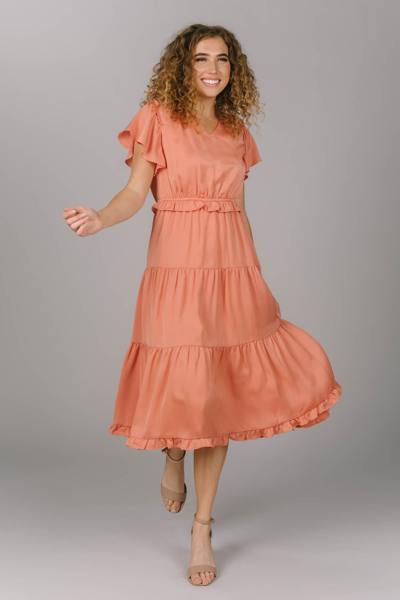 Front view of a peach modest bridesmaid dress. It has a v-neckline and flutter sleeves. Its draping and tiered skirt make for the perfect modest bridesmaid dress.