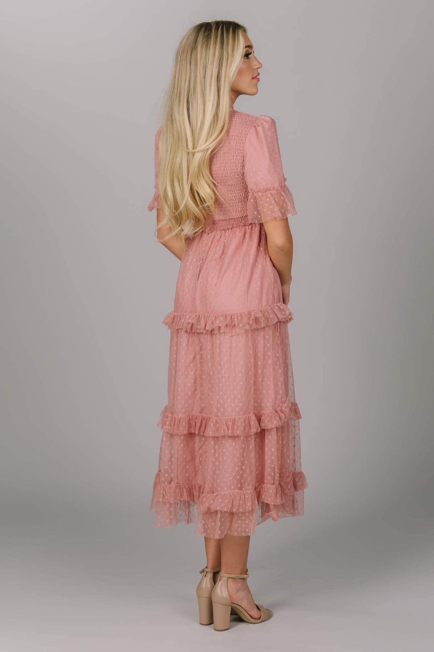 Back view of blush modest dress. It has ruffled tiering on the sleeves and skirt. This midi-length dress has a higher neckline and slightly puffed sleeves. The Pyper dress could make a fun modest bridesmaid dress.
