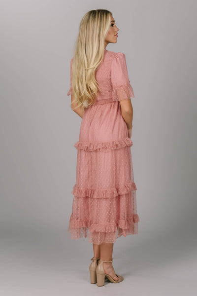Back view of blush modest dress. It has ruffled tiering on the sleeves and skirt. This midi-length dress has a higher neckline and slightly puffed sleeves. The Pyper dress could make a fun modest bridesmaid dress.